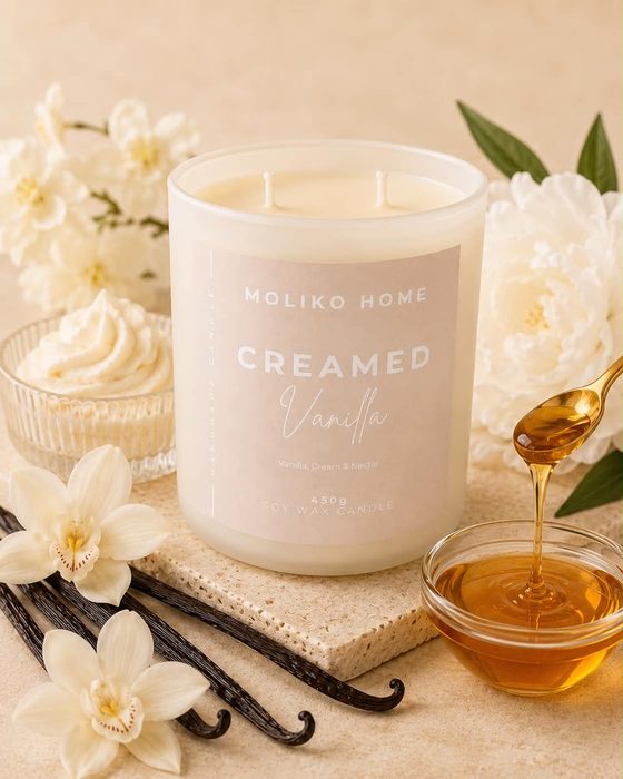 The Creamed Vanilla candle in its cream glass vessel with a pale greige label, beside vanilla flowers, whipped cream and honey.