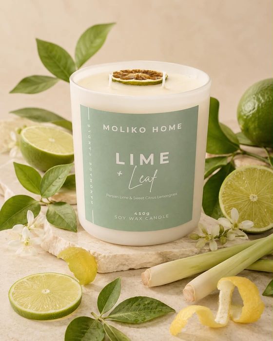 The Lime & Leaf candle in its cream glass vessel with a sage green label, topped with a dried lime slice, beside limes and lemongrass.
