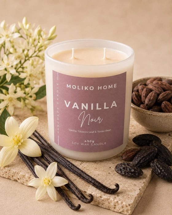 The Vanilla Noir candle in its cream glass vessel with a dusty mauve label, beside vanilla pods and cacao beans.